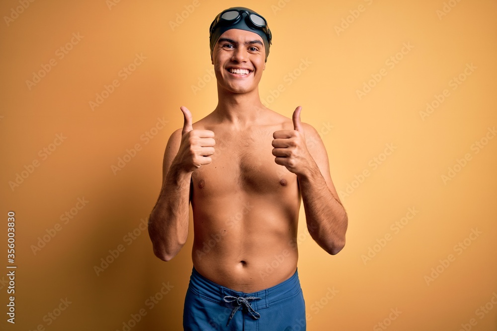 Young handsome man shirtless wearing swimsuit and swim cap over isolated yellow background success sign doing positive gesture with hand, thumbs up smiling and happy. Cheerful expression and winner