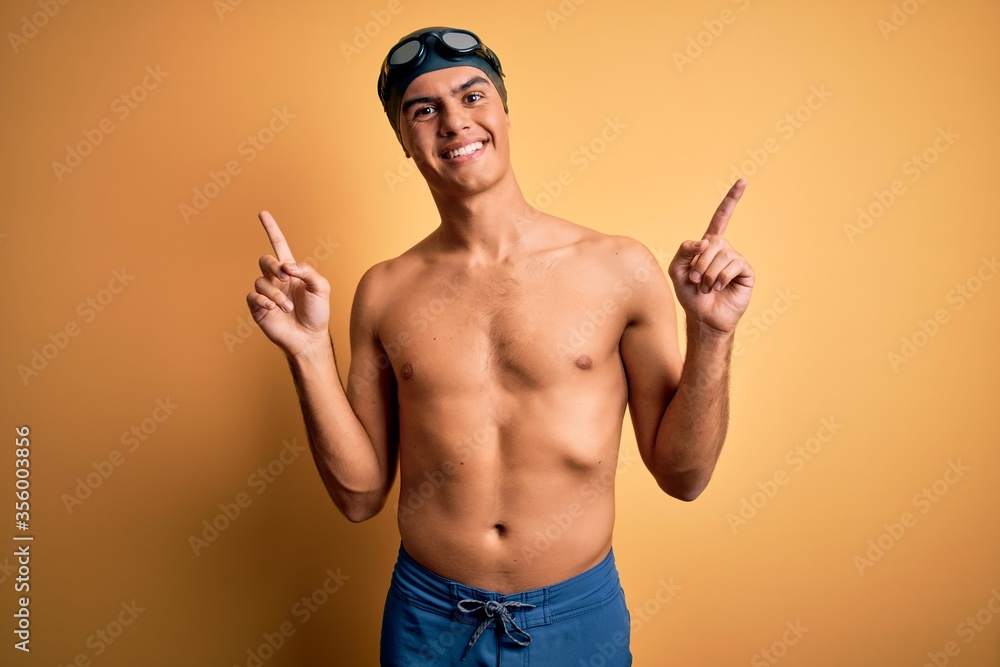 Young handsome man shirtless wearing swimsuit and swim cap over isolated yellow background smiling confident pointing with fingers to different directions. Copy space for advertisement