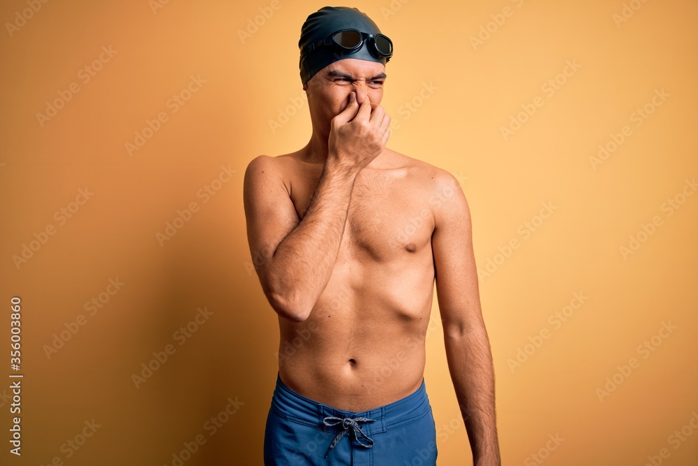 Young handsome man shirtless wearing swimsuit and swim cap over isolated yellow background smelling something stinky and disgusting, intolerable smell, holding breath with fingers on nose. Bad smell