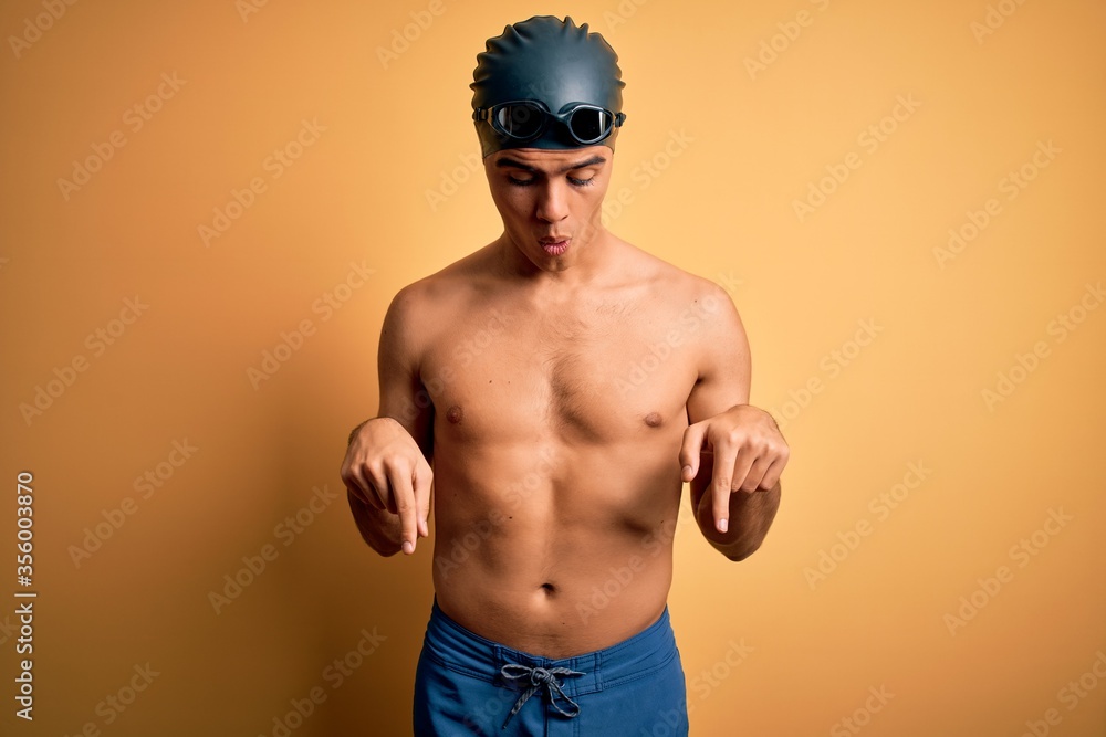 Young handsome man shirtless wearing swimsuit and swim cap over isolated yellow background Pointing down with fingers showing advertisement, surprised face and open mouth