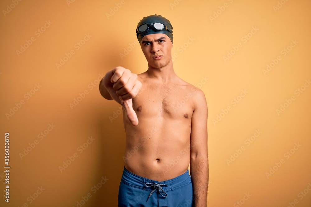 Young handsome man shirtless wearing swimsuit and swim cap over isolated yellow background looking unhappy and angry showing rejection and negative with thumbs down gesture. Bad expression.