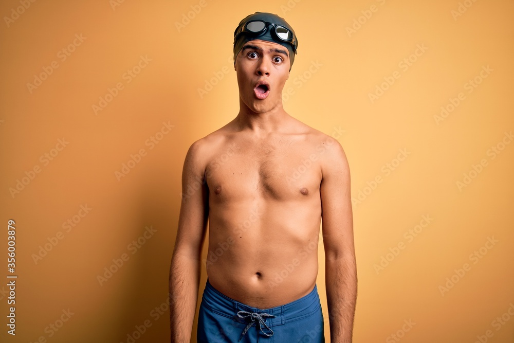 Young handsome man shirtless wearing swimsuit and swim cap over isolated yellow background In shock face, looking skeptical and sarcastic, surprised with open mouth