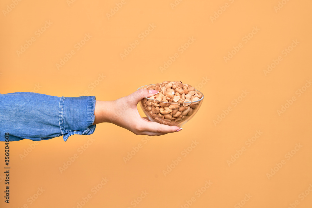 Obraz premium Beautiful hand of woman holding bowl with healthy salty peanuts over isolated yellow background