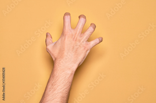 Hand of caucasian young man showing fingers over isolated yellow background grasping aggressive and scary with fingers  violence and frustration