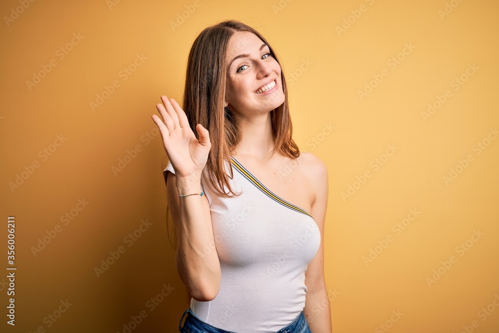 Young beautiful redhead woman wearing casual t-shirt over isolated yellow background Waiving saying hello happy and smiling, friendly welcome gesture Young beautiful redhead woman wearing casual t-shirt over isolated yellow background Waiving saying hello happy and smiling, friendly welcome gesture