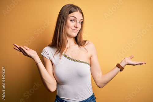 Young beautiful redhead woman wearing casual t-shirt over isolated yellow background clueless and confused expression with arms and hands raised. Doubt concept.
