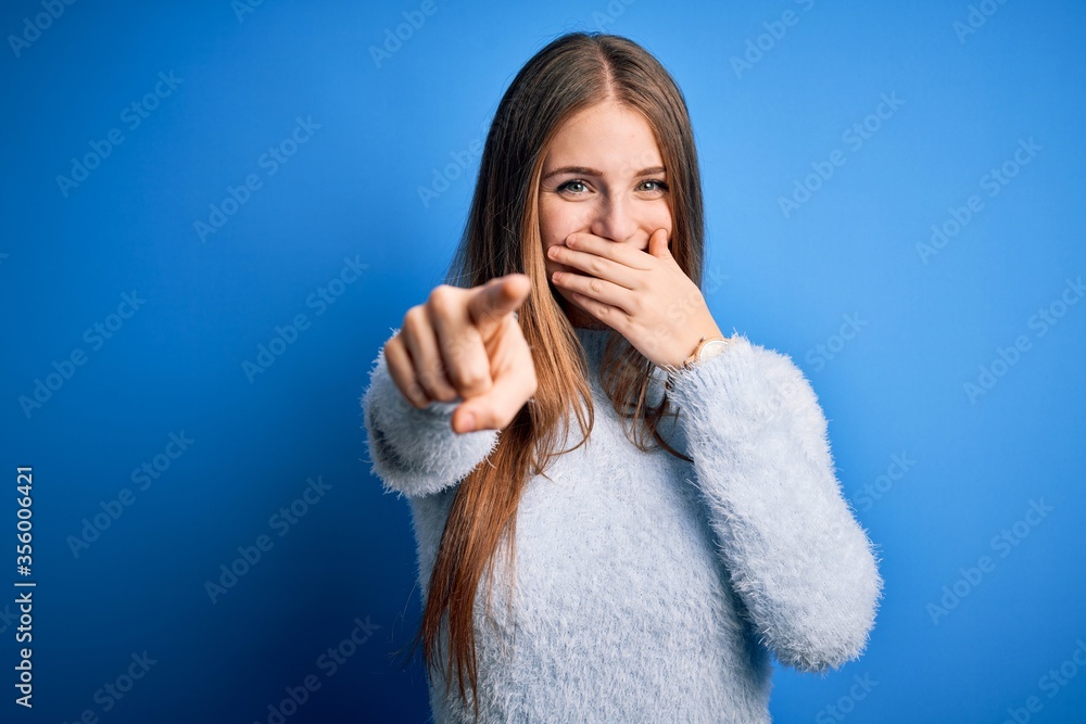 Young beautiful redhead woman wearing casual sweater over isolated blue background laughing at you, pointing finger to the camera with hand over mouth, shame expression Young beautiful redhead woman wearing casual sweater over isolated blue background laughing at you, pointing finger to the camera with hand over mouth, shame expression
