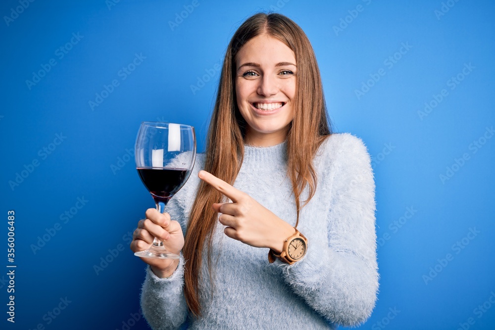 Young beautiful redhead woman drinking glass of red wine over isolated blue background very happy pointing with hand and finger Young beautiful redhead woman drinking glass of red wine over isolated blue background very happy pointing with hand and finger