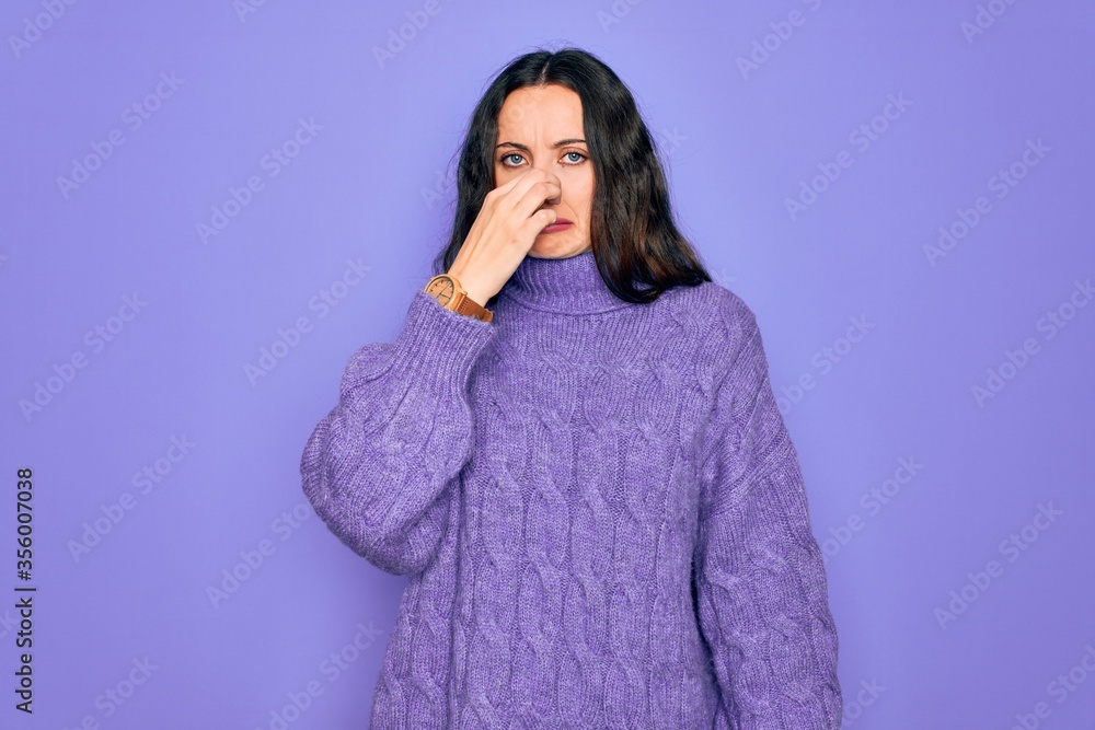 Young beautiful woman wearing casual turtleneck sweater standing over purple background smelling something stinky and disgusting, intolerable smell, holding breath with fingers on nose. Bad smell Young beautiful woman wearing casual turtleneck sweater standing over purple background smelling something stinky and disgusting, intolerable smell, holding breath with fingers on nose. Bad smell