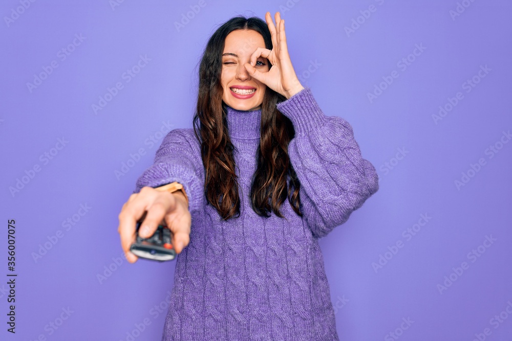 Young beautiful woman using television remote control over isolated purple background with happy face smiling doing ok sign with hand on eye looking through fingers Young beautiful woman using television remote control over isolated purple background with happy face smiling doing ok sign with hand on eye looking through fingers