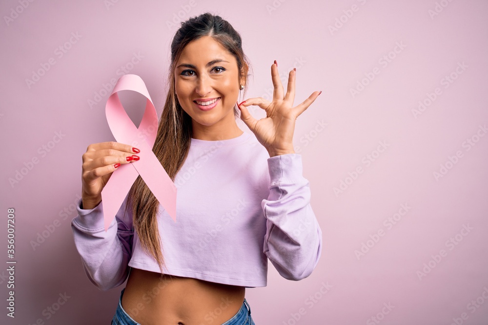 Young beautiful brunette woman holding pink cancer ribbon symbol over isolated background doing ok sign with fingers, excellent symbol Young beautiful brunette woman holding pink cancer ribbon symbol over isolated background doing ok sign with fingers, excellent symbol