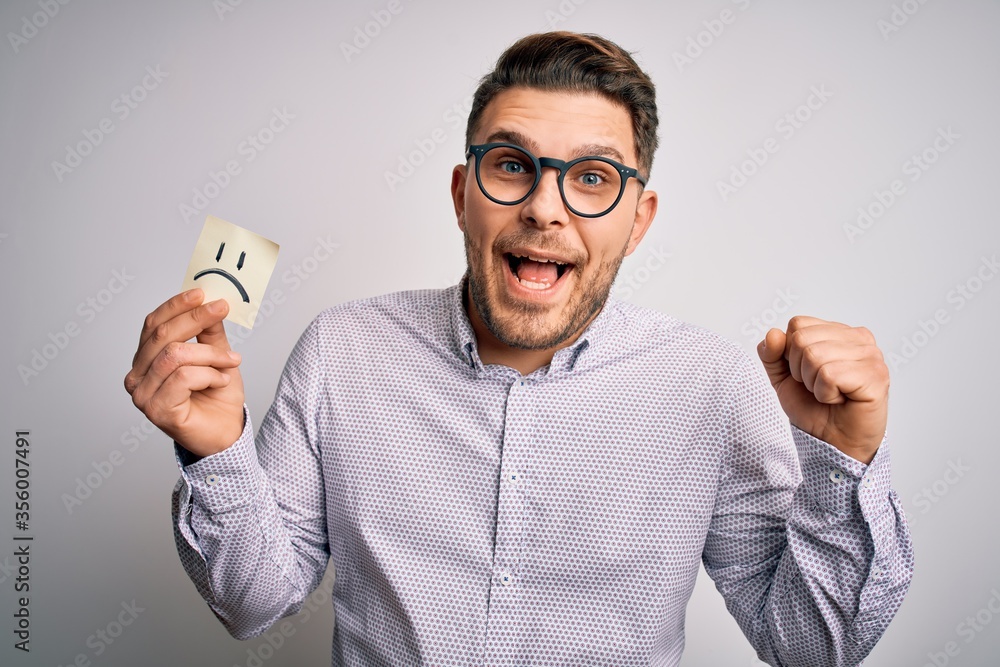 Young business man with blue eyes holding paper note with sad face as unhappy message screaming proud and celebrating victory and success very excited, cheering emotion Young business man with blue eyes holding paper note with sad face as unhappy message screaming proud and celebrating victory and success very excited, cheering emotion
