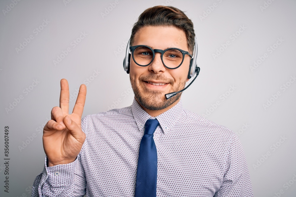 Young call center operator business man with blue eyes wearing glasses and headset smiling with happy face winking at the camera doing victory sign. Number two. Young call center operator business man with blue eyes wearing glasses and headset smiling with happy face winking at the camera doing victory sign. Number two.
