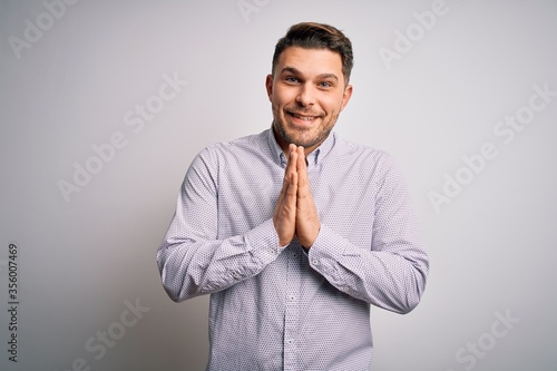 Young business man with blue eyes standing over isolated background praying with hands together asking for forgiveness smiling confident.