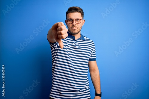 Young man with blue eyes wearing glasses and casual striped t-shirt over blue background looking unhappy and angry showing rejection and negative with thumbs down gesture. Bad expression.
