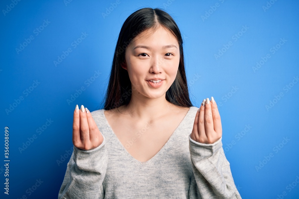 Young beautiful asian woman wearing casual sweater standing over blue isolated background doing money gesture with hands, asking for salary payment, millionaire business