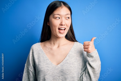 Young beautiful asian woman wearing casual sweater standing over blue isolated background smiling with happy face looking and pointing to the side with thumb up.