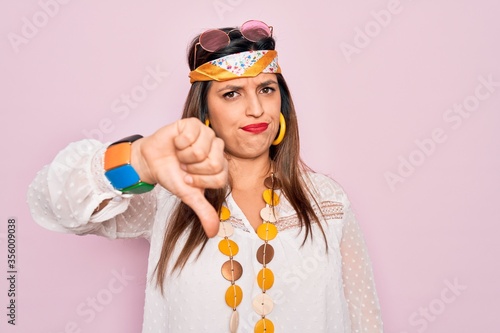 Young hispanic hippie woman wearing fashion boho style and sunglasses over pink background looking unhappy and angry showing rejection and negative with thumbs down gesture. Bad expression.