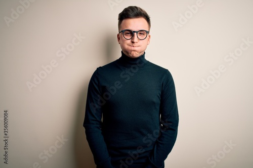Young handsome caucasian man wearing glasses and casual sweater over isolated background puffing cheeks with funny face. Mouth inflated with air, crazy expression. © Krakenimages.com