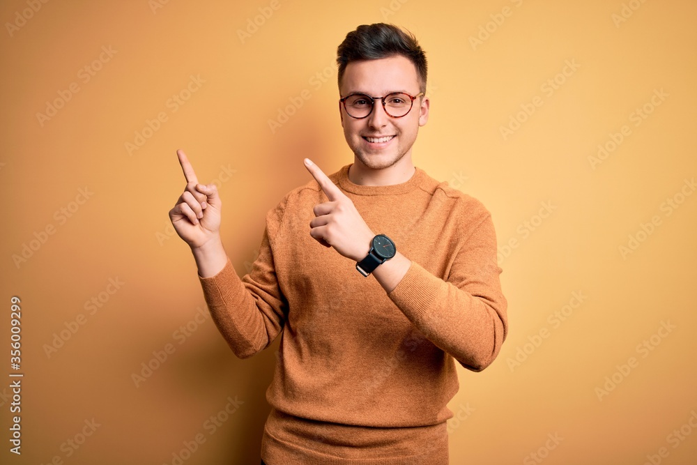 Young handsome caucasian man wearing glasses and casual winter sweater over yellow background smiling and looking at the camera pointing with two hands and fingers to the side.