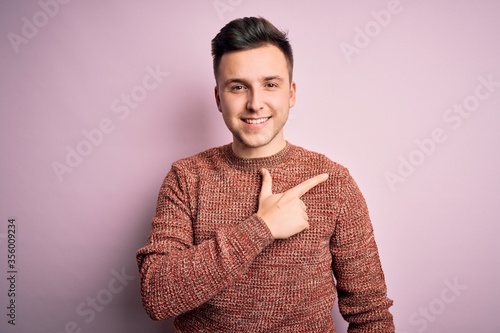 Young handsome caucasian man wearing casual winter sweater over pink isolated background cheerful with a smile on face pointing with hand and finger up to the side with happy and natural expression