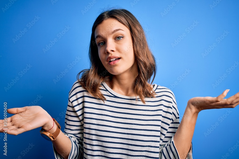 Fototapeta premium Young beautiful blonde girl wearing casual sweater standing over blue isolated background clueless and confused expression with arms and hands raised. Doubt concept.