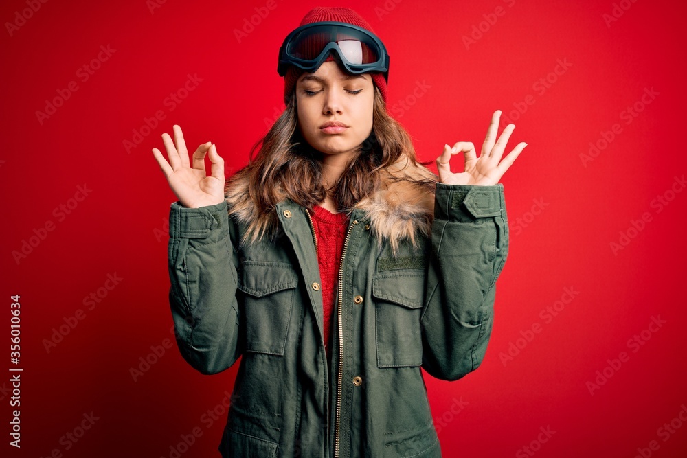 Young blonde girl wearing ski glasses and winter coat for ski weather over red background relaxed and smiling with eyes closed doing meditation gesture with fingers. Yoga concept.
