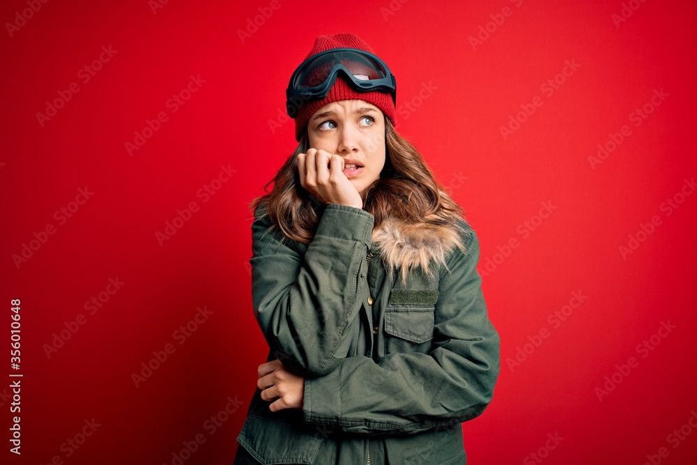 Young blonde girl wearing ski glasses and winter coat for ski weather over red background looking stressed and nervous with hands on mouth biting nails. Anxiety problem. Young blonde girl wearing ski glasses and winter coat for ski weather over red background looking stressed and nervous with hands on mouth biting nails. Anxiety problem.