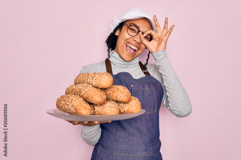 Young african american baker woman wearing apron holding wholemeal bread with cereal with happy face smiling doing ok sign with hand on eye looking through fingers Young african american baker woman wearing apron holding wholemeal bread with cereal with happy face smiling doing ok sign with hand on eye looking through fingers