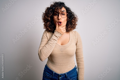 Young beautiful curly arab woman wearing casual t-shirt and glasses over white background Looking fascinated with disbelief, surprise and amazed expression with hands on chin