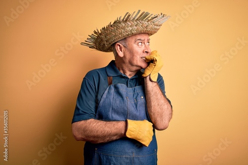 Middle age hoary farmer man wearing apron and hat over isolated yellow background looking stressed and nervous with hands on mouth biting nails. Anxiety problem.