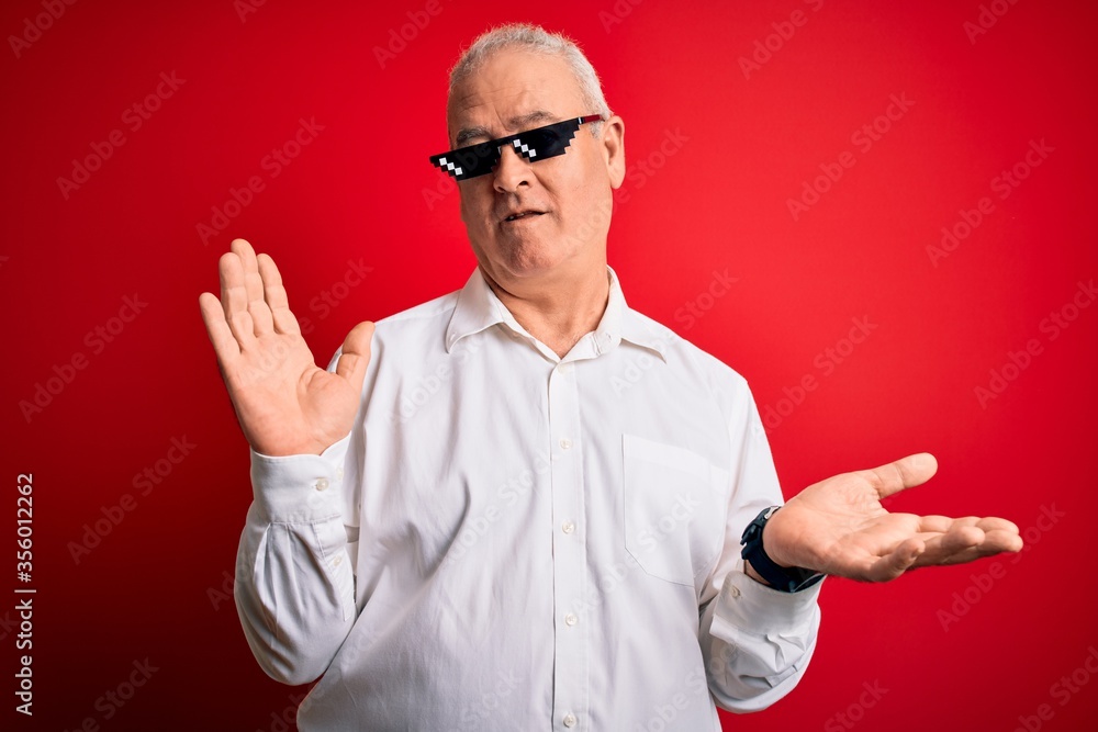 Naklejka premium Middle age hoary man wearing funny sunglasses over isolated red background clueless and confused expression with arms and hands raised. Doubt concept.