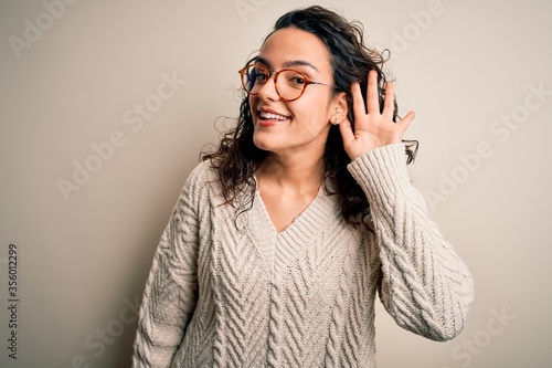 Beautiful woman with curly hair wearing casual sweater and glasses over white background smiling with hand over ear listening an hearing to rumor or gossip. Deafness concept.