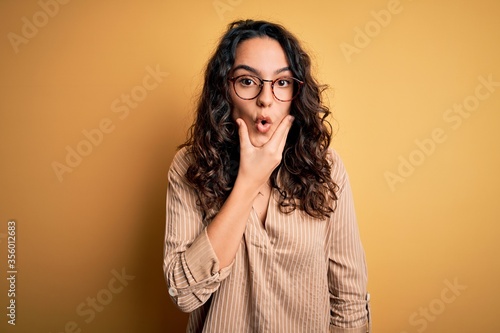 Beautiful woman with curly hair wearing striped shirt and glasses over yellow background Looking fascinated with disbelief, surprise and amazed expression with hands on chin