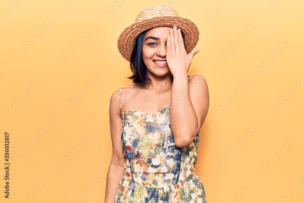 Young beautiful latin woman wearing summer hat covering one eye with hand, confident smile on face and surprise emotion. Young beautiful latin woman wearing summer hat covering one eye with hand, confident smile on face and surprise emotion.
