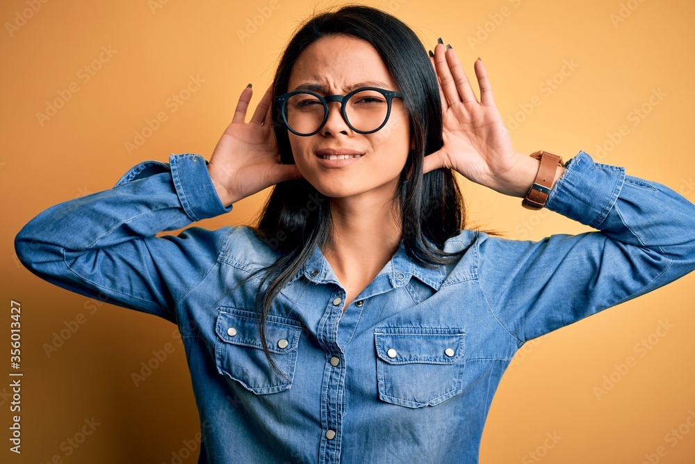 Young beautiful chinese woman wearing casual denim shirt over isolated yellow background Trying to hear both hands on ear gesture, curious for gossip. Hearing problem, deaf