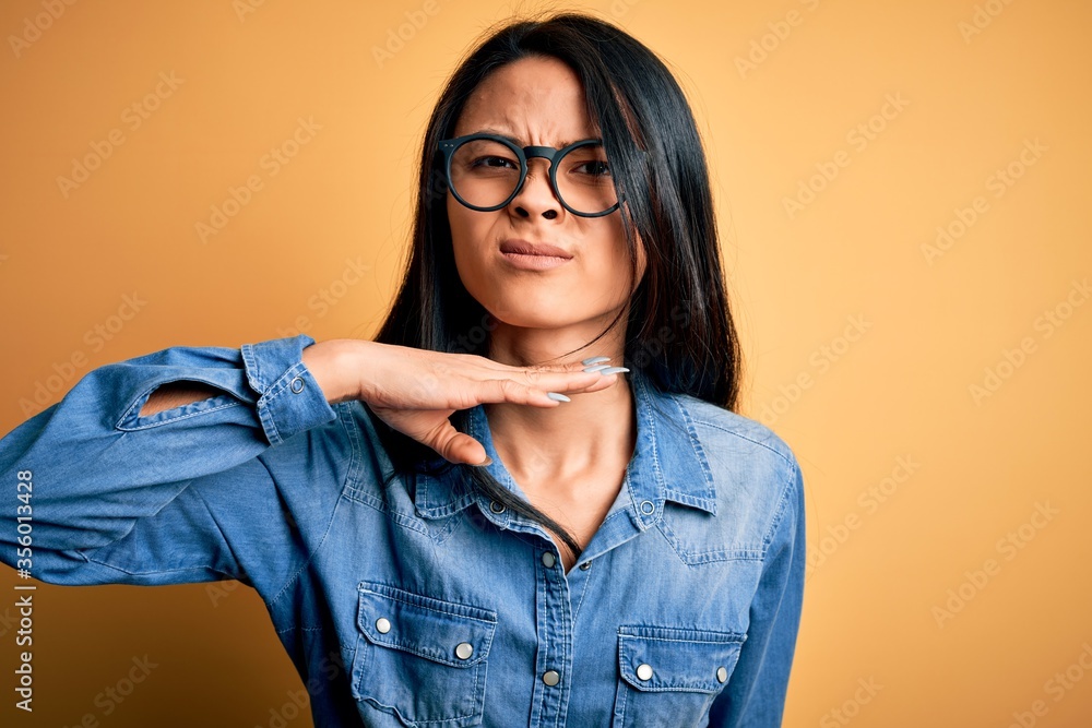 Young beautiful chinese woman wearing casual denim shirt over isolated yellow background cutting throat with hand as knife, threaten aggression with furious violence