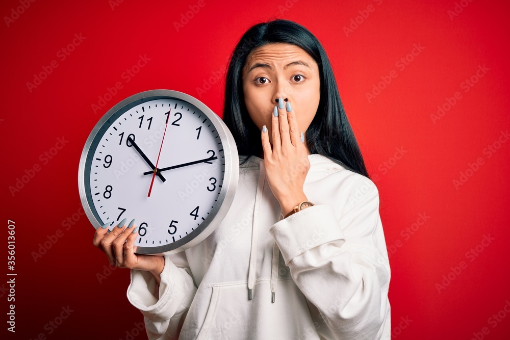 Young beautiful chinese woman holding big clock standing over isolated red background cover mouth with hand shocked with shame for mistake, expression of fear, scared in silence, secret concept Young beautiful chinese woman holding big clock standing over isolated red background cover mouth with hand shocked with shame for mistake, expression of fear, scared in silence, secret concept