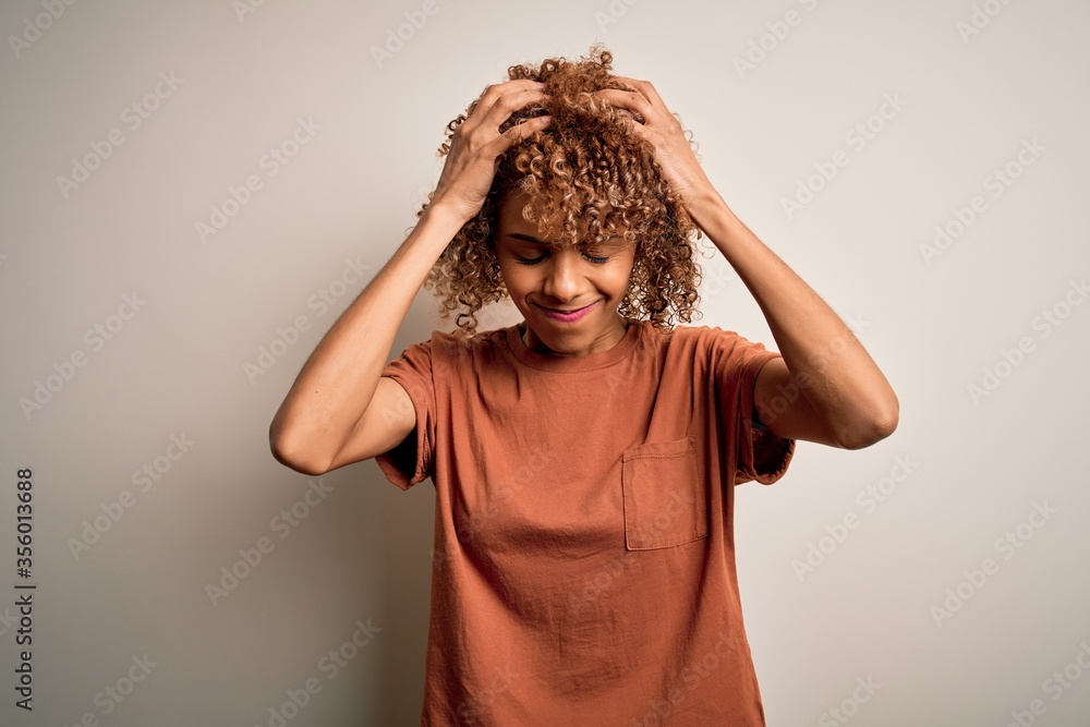Beautiful african american woman with curly hair wearing casual t-shirt over white background suffering from headache desperate and stressed because pain and migraine. Hands on head. Beautiful african american woman with curly hair wearing casual t-shirt over white background suffering from headache desperate and stressed because pain and migraine. Hands on head.