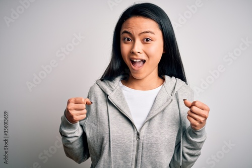 Young beautiful chinese sporty woman wearing sweatshirt over isolated white background celebrating surprised and amazed for success with arms raised and open eyes. Winner concept. © Krakenimages.com