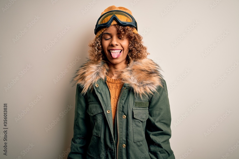 Young african american skier woman with curly hair wearing snow sportswear and ski goggles sticking tongue out happy with funny expression. Emotion concept.