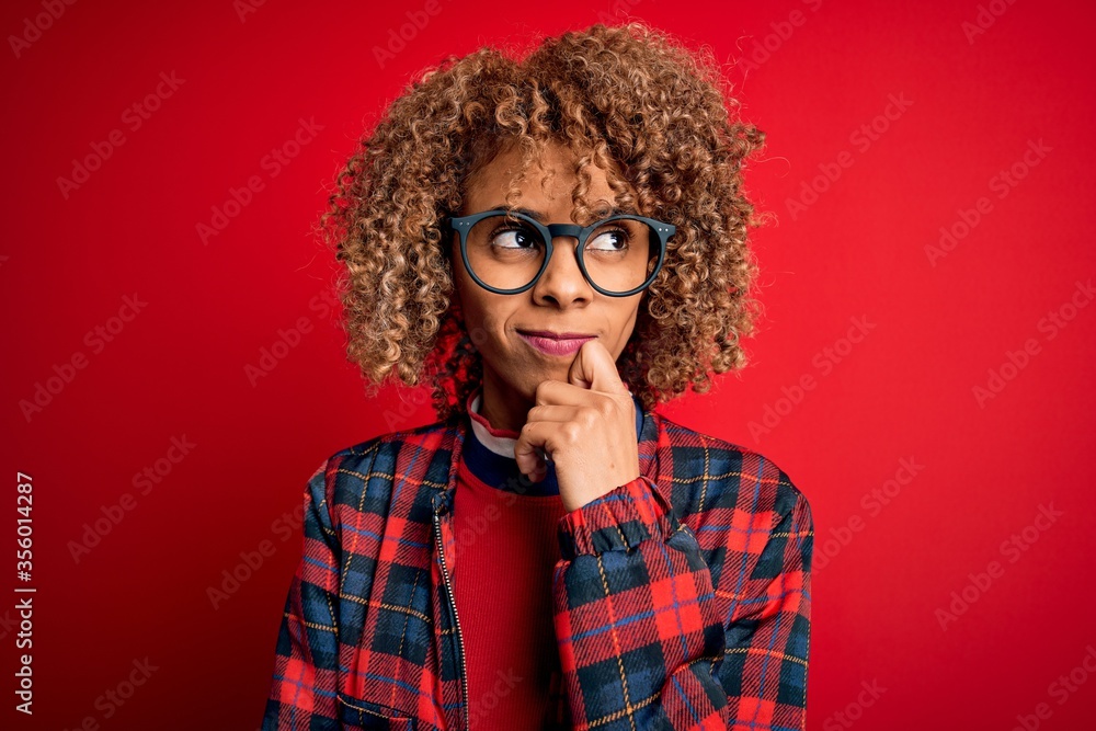 Young beautiful african american woman wearing casual shirt and glasses over red background with hand on chin thinking about question, pensive expression. Smiling with thoughtful face. Doubt concept. Young beautiful african american woman wearing casual shirt and glasses over red background with hand on chin thinking about question, pensive expression. Smiling with thoughtful face. Doubt concept.