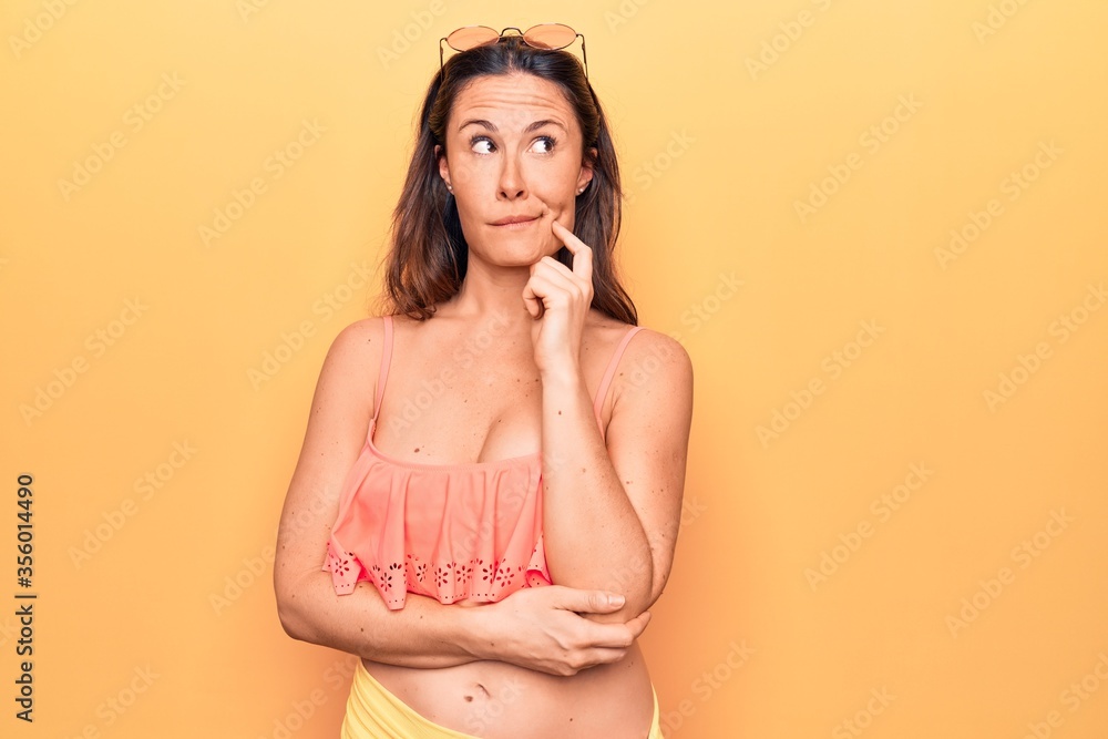Obraz premium Young beautiful brunette woman wearing bikini standing over isolated yellow background thinking concentrated about doubt with finger on chin and looking up wondering