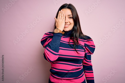 Young brunette elegant woman wearing striped shirt over pink isolated background covering one eye with hand, confident smile on face and surprise emotion. © Krakenimages.com