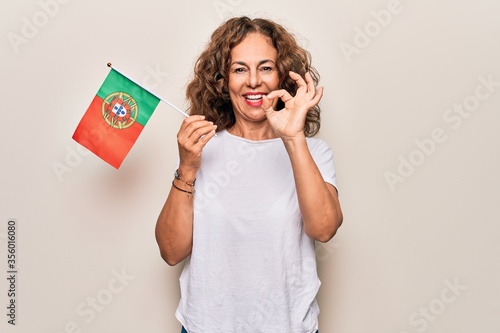 Middle age beautiful patriotic woman holding portuguese flag over isolated white background doing ok sign with fingers, smiling friendly gesturing excellent symbol