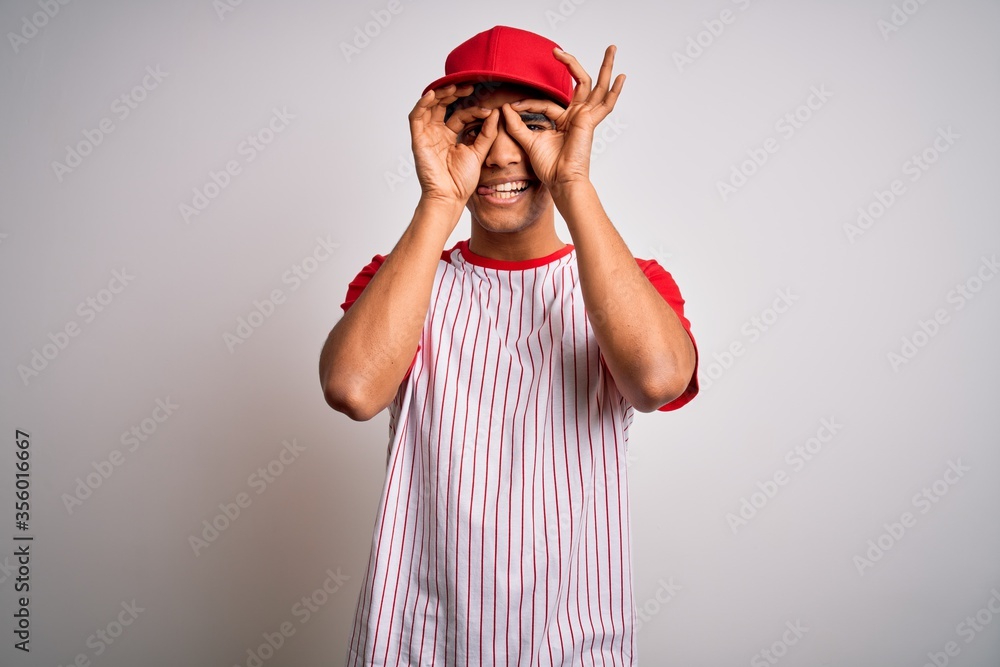 Young handsome african american sportsman wearing striped baseball t-shirt and cap doing ok gesture like binoculars sticking tongue out, eyes looking through fingers. Crazy expression.