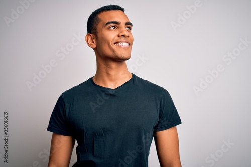 Young handsome african american man wearing casual t-shirt standing over white background looking away to side with smile on face, natural expression. Laughing confident.
