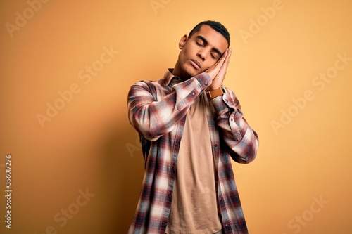 Young handsome african american man wearing casual shirt standing over yellow background sleeping tired dreaming and posing with hands together while smiling with closed eyes.