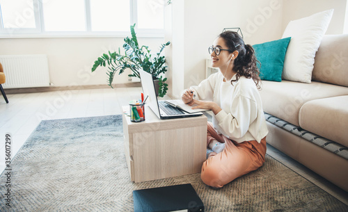 Smart caucasian girl with eyeglasses sitting on floor with a laptop while smile and having online lesson from home