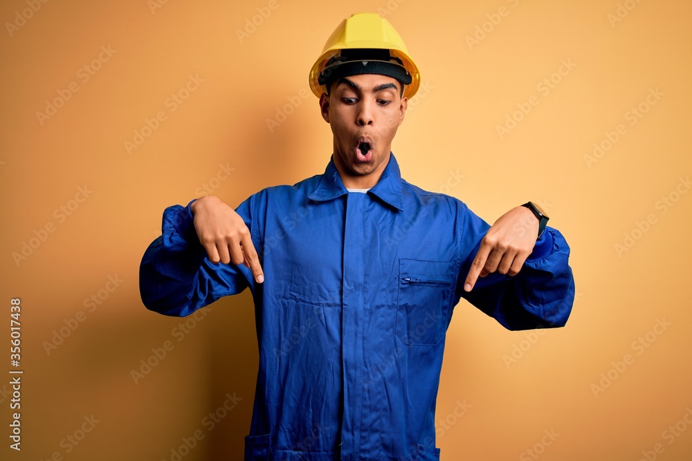 Young handsome african american worker man wearing blue uniform and security helmet Pointing down with fingers showing advertisement, surprised face and open mouth